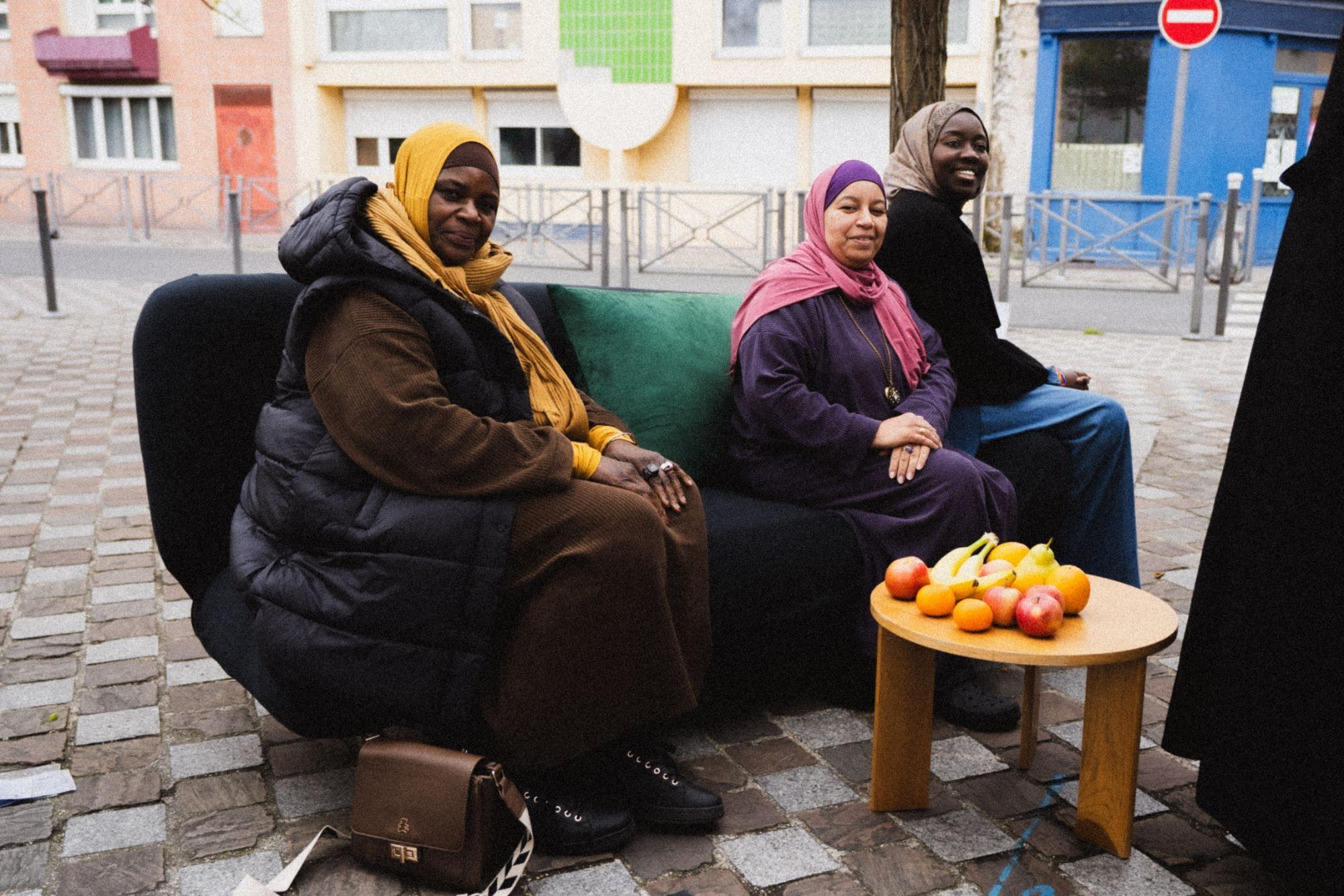 Sur une place pavée, trois femmes voilées assises sur un canapé noir installé dans la rue sourient à l'objectif ; devant elles, une table basse en bois garnie de fruits ; derrière, des façades colorées.